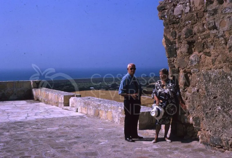 Two older people stand on a stone-paved terrace beside a rough stone wall, with fields and sea behind