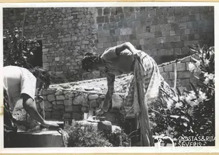 Two people bending over a stone basin with running water beside a rough stone wall and oleanders