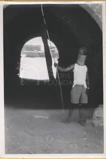 Man in a cylindrical helmet and white vest holding a pole with a dark flag, standing under a stone archway