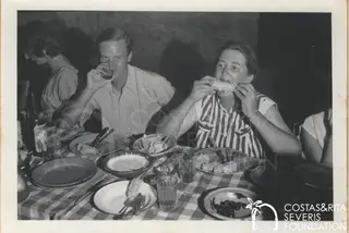 A man drinks from a glass and a woman in a striped top eats corn on the cob at a checked-cloth dinner table