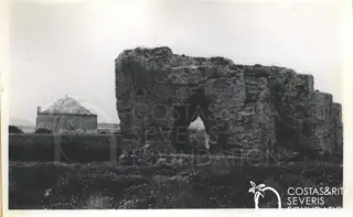 Ruined stone wall with an arched opening in a flowery field, with a domed building behind on the left