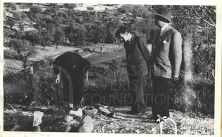 Woman bending over stones on a hillside, with two men in suits and hats standing beside her