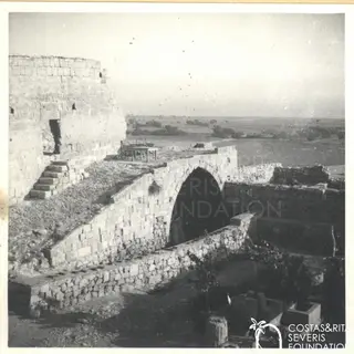 Stone ruins with an arch, steps and a wooden table on a terrace, with open fields beyond