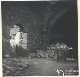 Vaulted stone chamber with sacks and cut stone blocks on the floor and a table of fragments at right