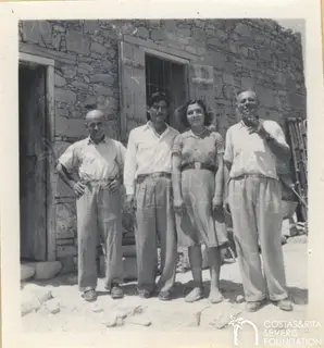 Four people stand side by side in front of a stone building with a wooden door and barred window