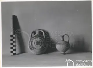Two clay vessels on a surface beside a checkered scale rod against a grey background