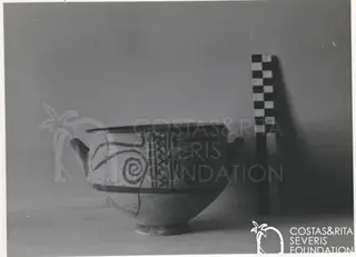 Footed pottery bowl with two side handles and dark painted spiral motifs, beside a chequered scale rod