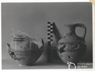 Two broken pottery vessels beside a chequered scale rod against a grey backdrop