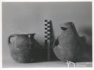 Two broken pottery vessels, a handled jug on the left and a larger jar on the right, with a chequered scale rod between them