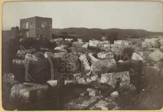 Scattered cut stone blocks among scrub, with a stone building at left and hills behind