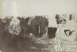 Rock-cut limestone cliff face with dark tomb openings and arched niches above rubble ground