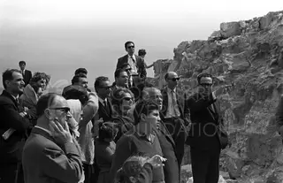 Group of visitors in suits and sunglasses gathered beside rough stone ruins, one man pointing ahead