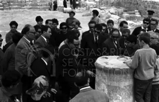 Crowd of men in suits and sunglasses gathered around a stone column drum, with a boy in a sweater at right