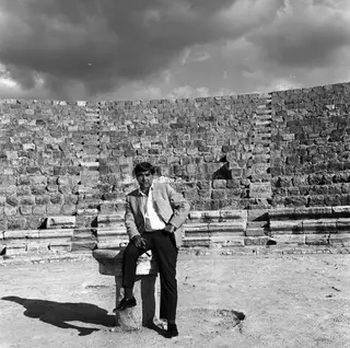 Man in a striped jacket sits on a stone block before a tall stone wall at Salamis under a cloudy sky