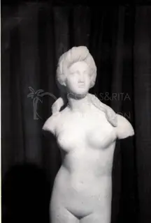 Marble statue of a female figure with a draped hairstyle and damaged arms, against a dark drape