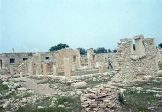 Ruined stone walls and column stumps on a grassy site, with a man walking among them