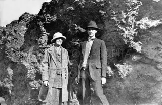 A woman and a man in hats stand before a craggy dark rock face