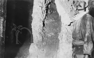 Man in a headcloth and loose shirt standing beside a rough rock wall inside a dark passage