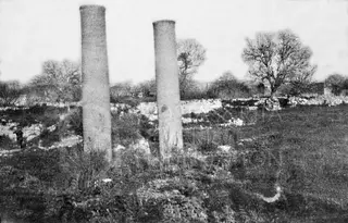 Two tall stone pillars standing in a grassy field among low ruined walls, with bare trees behind