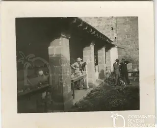 Woman at a table of pottery under a stone arcade, with two men in suits and hats standing nearby