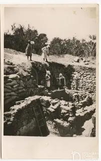 Workers dig in a deep excavation trench among stone walls, with figures standing on the rim and a ladder on the left.