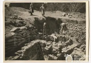 Workers excavating a trench with exposed stone walls, sandbags at left and women standing on the bank above
