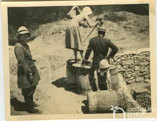 Four people by barrels and a stone wall on a dirt slope, one woman raising an arm as if taking a photograph