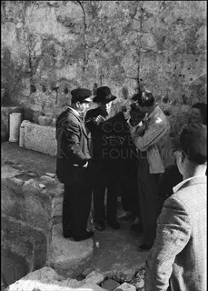 Men gathered in front of a rough stone wall, one in a dark hat speaking to the others