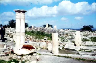 Ruined marble columns and a broken stone basin among scattered masonry, with a man on steps at left