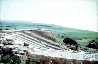 Curved stone tiers of a theatre on a hillside, with green fields and the coast beyond