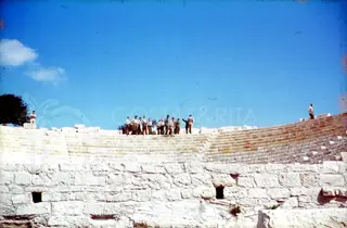 Group of men in uniform standing on the upper tiers of the stone seating of Kourion Theatre under a blue sky