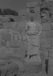 A man's head and shoulders rise behind a headless draped marble statue against a stone block wall