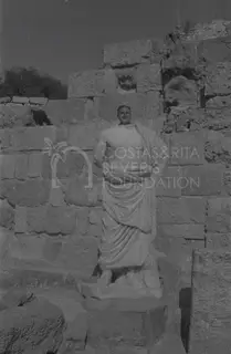 A man poses behind a draped marble statue in front of a stone block wall