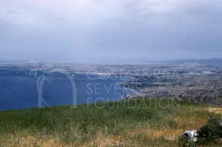Blue bay curving along a coast, with grassy ground in the foreground and hills and a settlement beyond