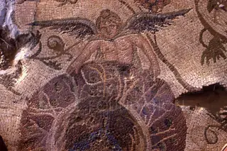 Close view of a mosaic floor showing a winged figure with a face, set among small coloured tesserae