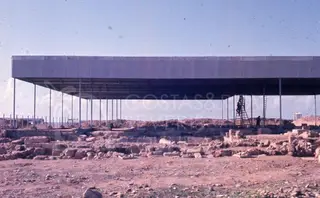 Steel canopy roof over stone wall remains at an excavated site, with figures beside a ladder at right