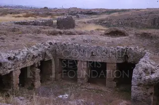 Rock-cut tomb facade with pillars and dark chamber openings in a dry, stony landscape