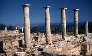 Tall stone columns stand above low walls, with trees and the sea beyond at Kourion