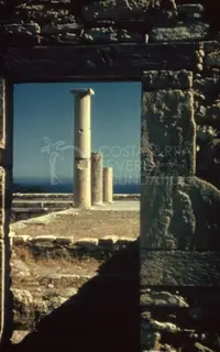 Three stone columns seen through a gap in a stone wall, with the sea on the horizon
