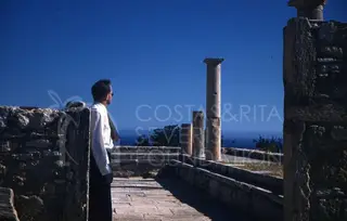 Man in white shirt stands on paved path among ancient stone walls and columns, facing the sea