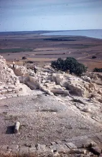 Two men stand among stone ruins and walls on a hilltop, with the flat coastal plain and sea beyond.