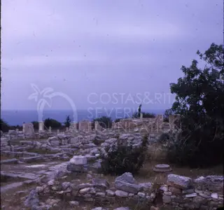 Ruined stone walls and scattered blocks of an ancient site, with the sea on the horizon and a figure standing among the ruins