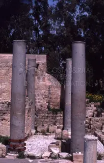 Grey columns stand above a geometric mosaic floor, with a brick wall and dark trees behind them