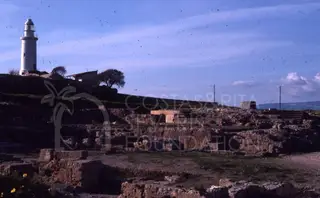 White lighthouse on a hill at left, above stone wall remains and rubble in the foreground