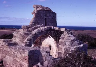 Ruined stone church with an arched opening and a broken tower, with the sea behind it