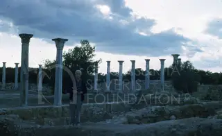 An elderly man stands in front of a row of tall columns at Salamis under a cloudy sky
