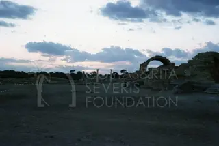 Ruins at dusk with a rounded arch on the right and standing columns in the middle distance under a cloudy sky