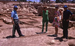 Four people in hats and sunglasses stand in an excavated site at Kition, with workers and stone walls behind them