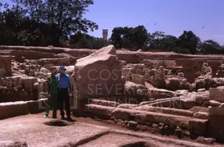 Two people stand among excavated stone ruins with column bases and a large fallen stone block at Kition.