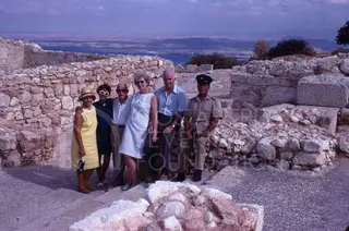 Five people and a uniformed guard posing among stone ruins with the sea behind
